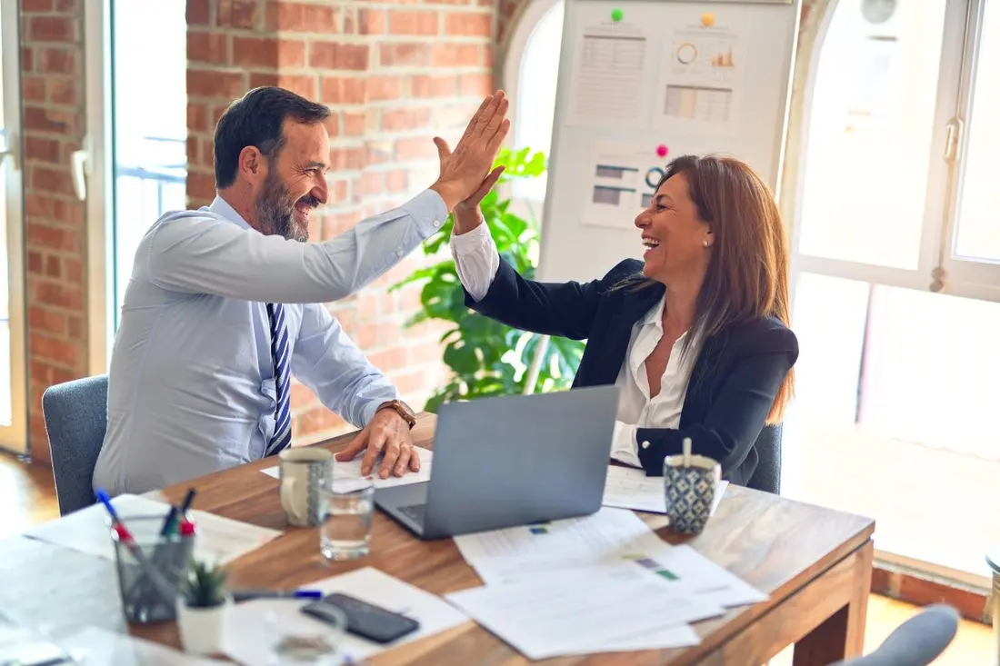 Two business professionals high-five in a modern office celebrating teamwork success