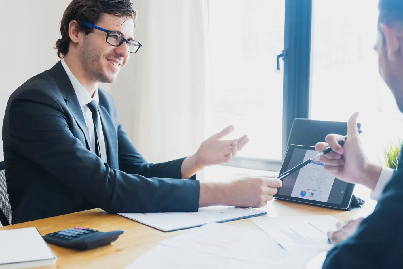 Two businessmen in formal attire discussing a business report in an office setting with a tablet