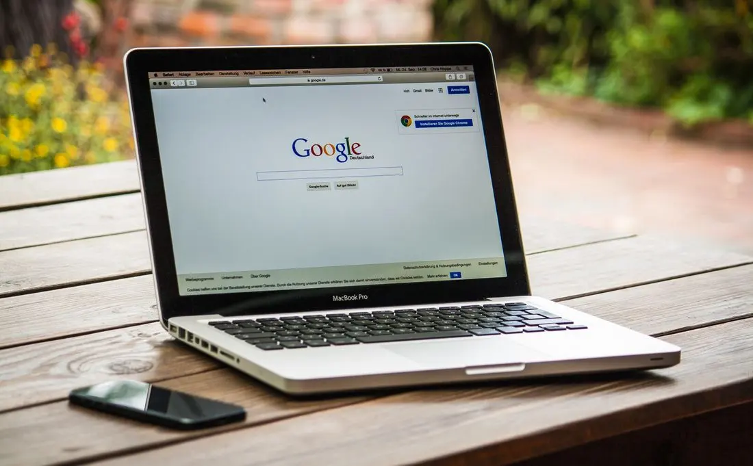 A MacBook Pro displaying Google Search on a wooden table outdoors, next to a smartphone