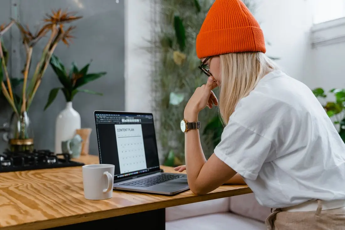 Young woman in orange beanie working on a laptop with a content plan in a cozy home office setting