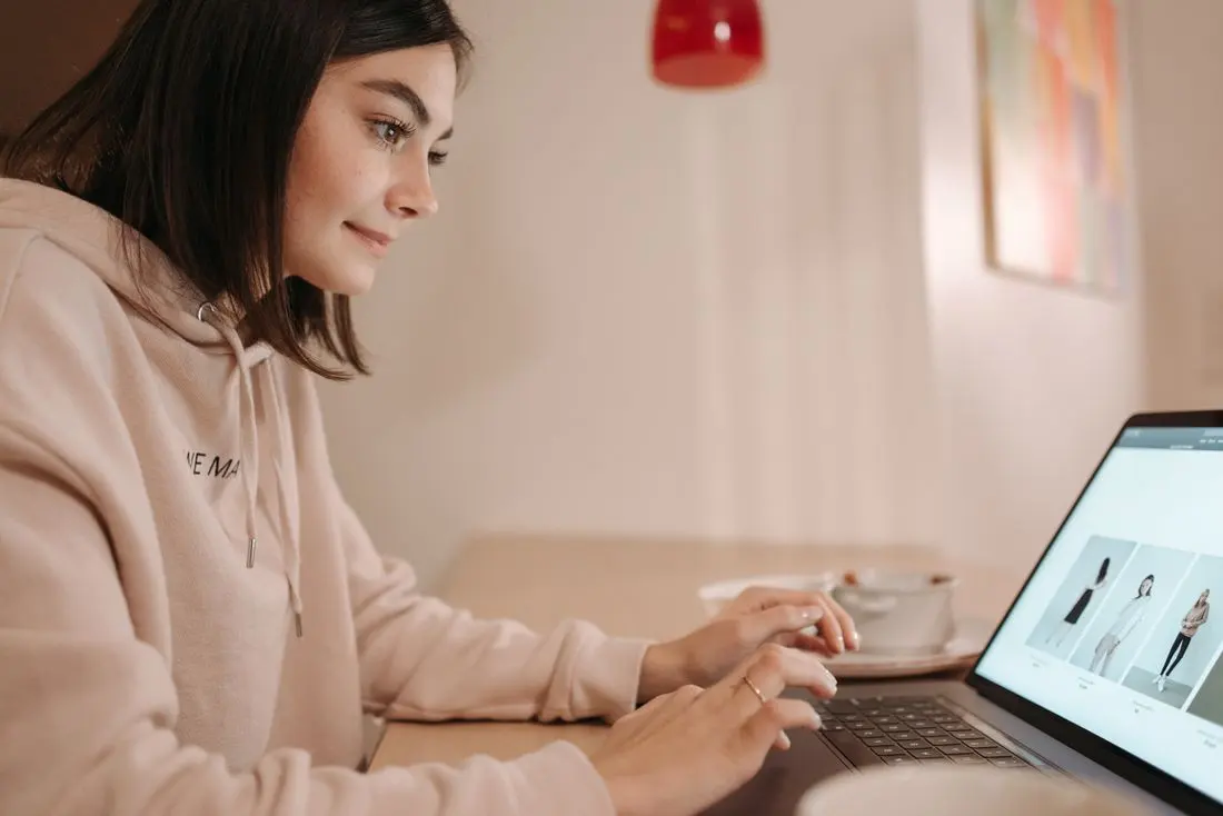 Young woman in pink hoodie browsing online fashion store on laptop at home. Calm and focused atmosphere